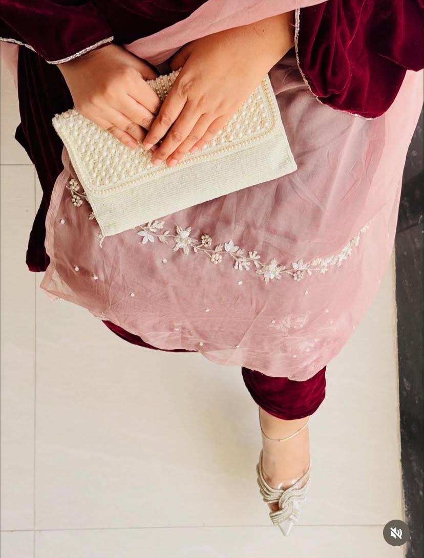A person wearing a dusty pink embroidered salwar kameez outfit with deep maroon velvet sleeves and holding a cream-colored pearl-embellished clutch purse, standing on a light tiled floor.