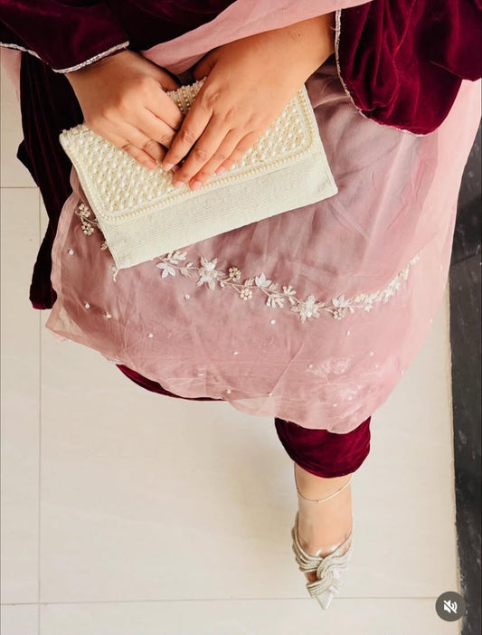 A person wearing a dusty pink embroidered salwar kameez outfit with deep maroon velvet sleeves and holding a cream-colored pearl-embellished clutch purse, standing on a light tiled floor.
