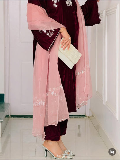 A person wearing a traditional outfit with a deep burgundy velvet kurta and matching trousers, paired with a light pink sheer dupatta featuring white floral embroidery. The person is holding a white clutch and wearing white heeled sandals in an indoor setting with white paneled walls.