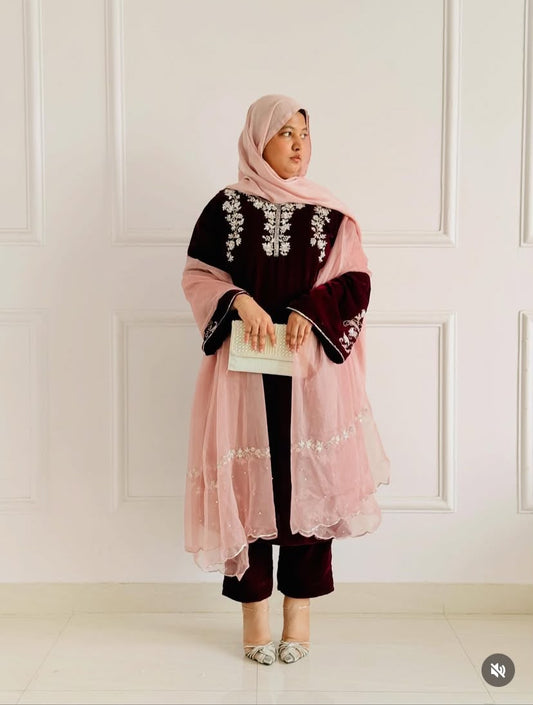 A woman modeling a traditional modest outfit featuring a dusty pink sheer tunic with silver embroidery over a deep maroon velvet tunic, matching maroon pants, and a light pink hijab, standing against a white paneled wall.