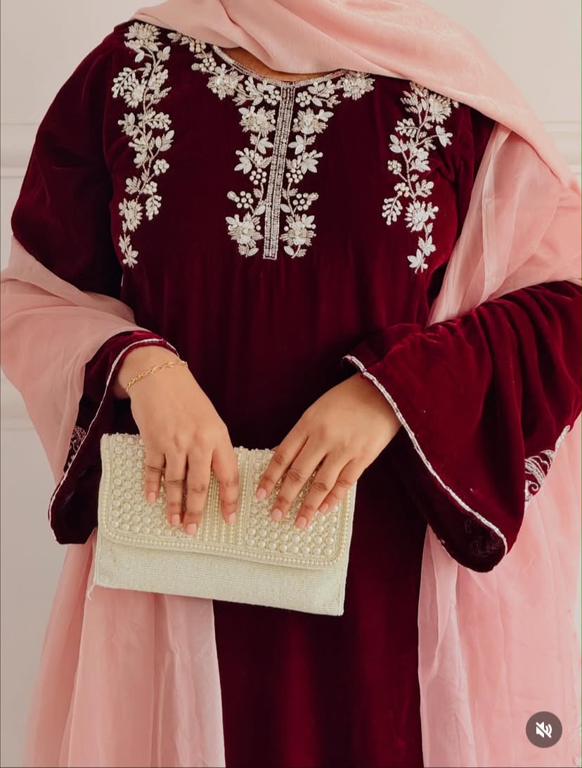 A person wearing a deep maroon velvet traditional outfit with white floral embroidery on the neckline, paired with a light pink dupatta and holding a white beaded clutch.