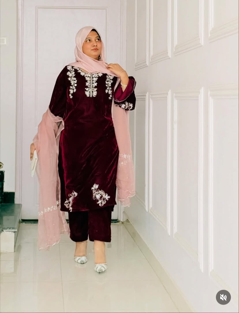 A woman wearing a traditional Islamic outfit, featuring a deep maroon velvet tunic with silver embroidery, matching maroon pants, a light pink hijab, and a sheer light pink dupatta, standing in a white paneled hallway.