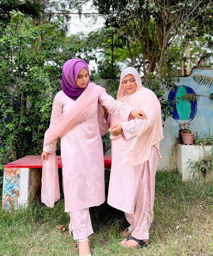 Two women wearing matching light pink traditional outfits and hijabs standing outdoors in a garden setting. One wears a purple hijab and the other wears a white hijab.