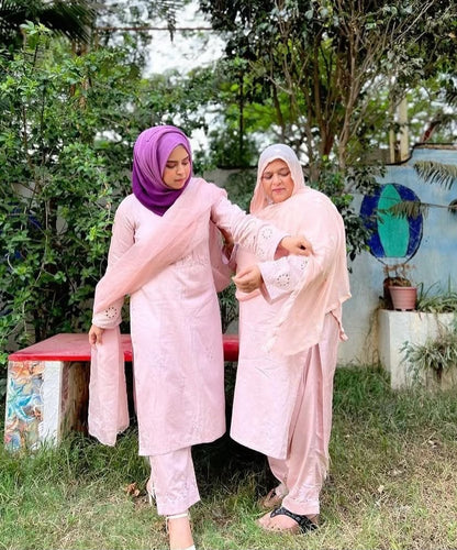 Two women wearing matching light pink traditional outfits and hijabs standing outdoors in a garden setting. One wears a purple hijab and the other wears a white hijab.