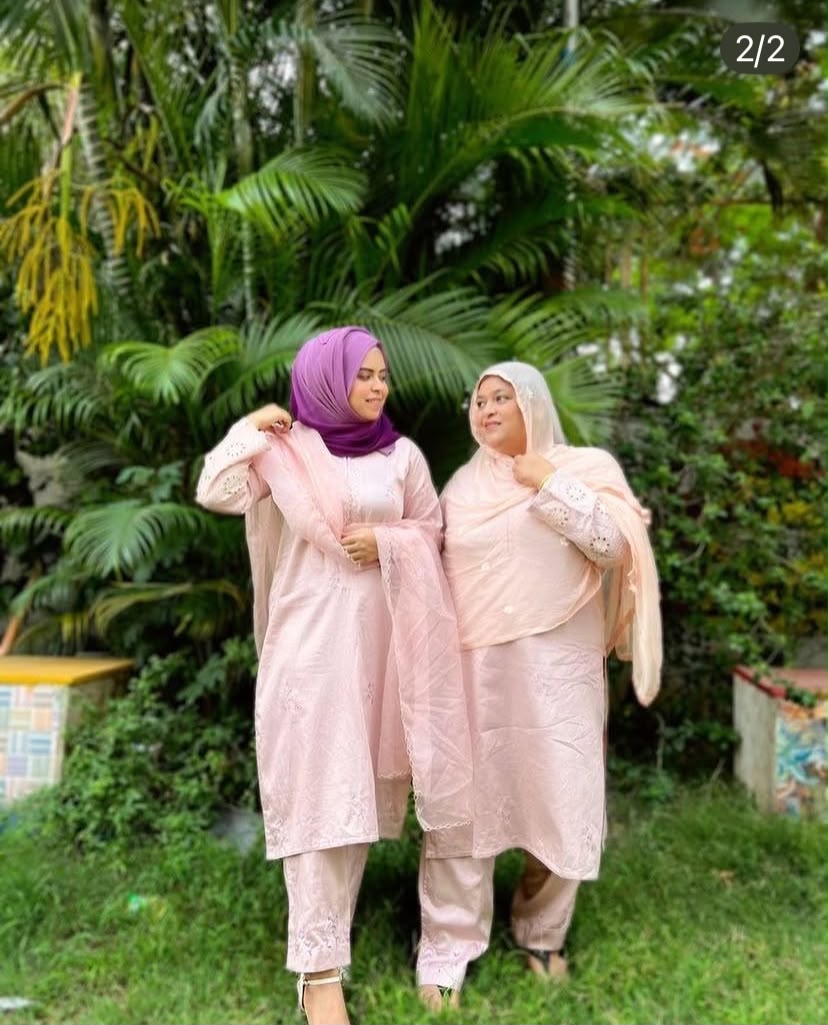 Two women wearing matching light pink traditional outfits with dupattas, one in a purple hijab and the other in a white hijab, standing outdoors in front of lush green tropical foliage.