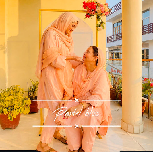 Two women wearing matching peach-pink traditional outfits and headscarves pose together on a sunlit balcony. One stands while the other kneels beside her, with a floral arch and building columns in the background.