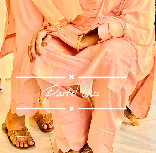 A person seated indoors wearing a pastel pink traditional outfit with matching pants and sandals, with gold bangles visible on the hands. White overlaid text reads 'Pastel pink'.