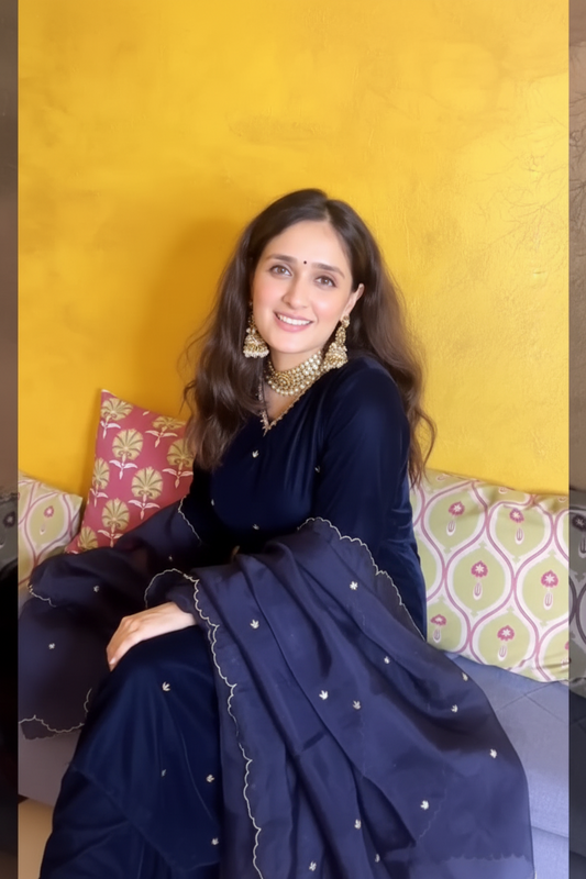 A woman wearing a navy blue traditional outfit with gold embellishments, seated on a sofa against a textured yellow wall.